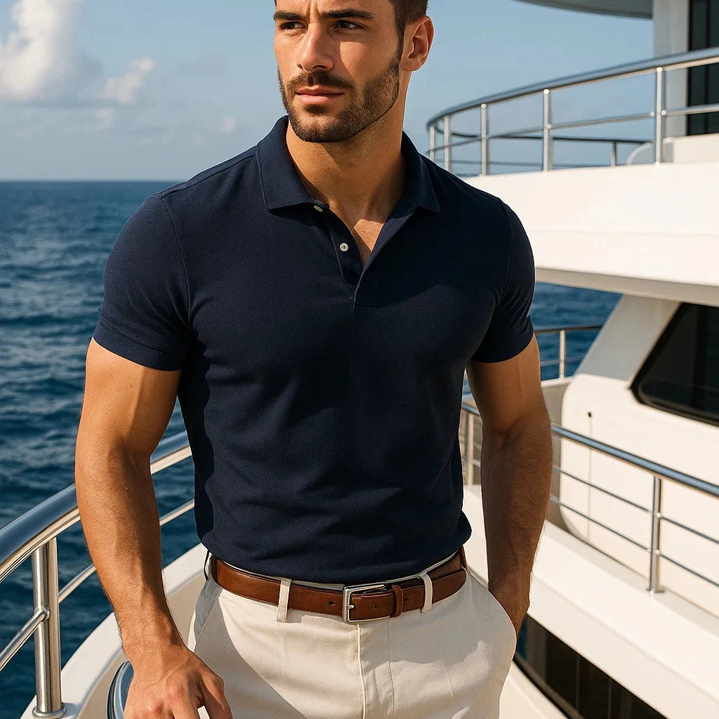 Man in navy polo shirt and beige pants standing on a yacht with ocean in the background